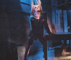 Liv Morgan dramatically flips her long blonde hair while leaning on a table in a black blazer and heels, shot in moody blue lighting for PAPER Magazine.