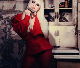 Liv Morgan poses powerfully in a red peplum blazer and tights, holding a knuckle duster near her face against a rusty industrial backdrop for PAPER Magazine.